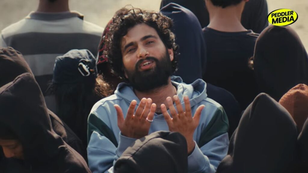 MC Square candid crowd moment wearing a light grey hoodie with curly hair and beard, pointing while talking in cinematic outdoor lighting with blurred people in background