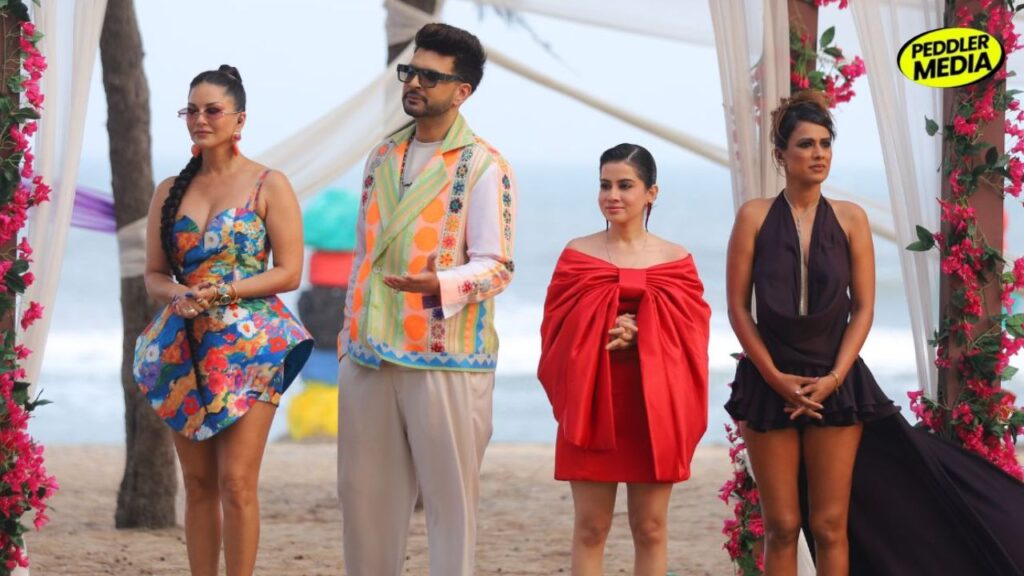 Four hosts of MTV Splitsvilla 16 stand together at a beachside set decorated with pink flowers and white drapes. Sunny Leone wears a colorful floral dress, Karan Kundrra sports a pastel embroidered jacket, Uorfi Javed appears in a bold red outfit, and Nia Sharma stuns in a dark flowing dress during the grand finale setup.