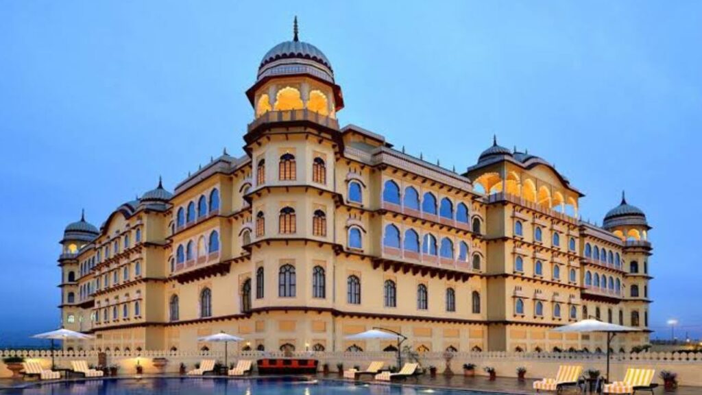 Grand heritage palace hotel architecture with illuminated arches next to a luxury outdoor swimming pool during twilight in Haryana in monsoon.
