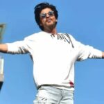 Shah Rukh Khan posing with his arms open against a blue sky