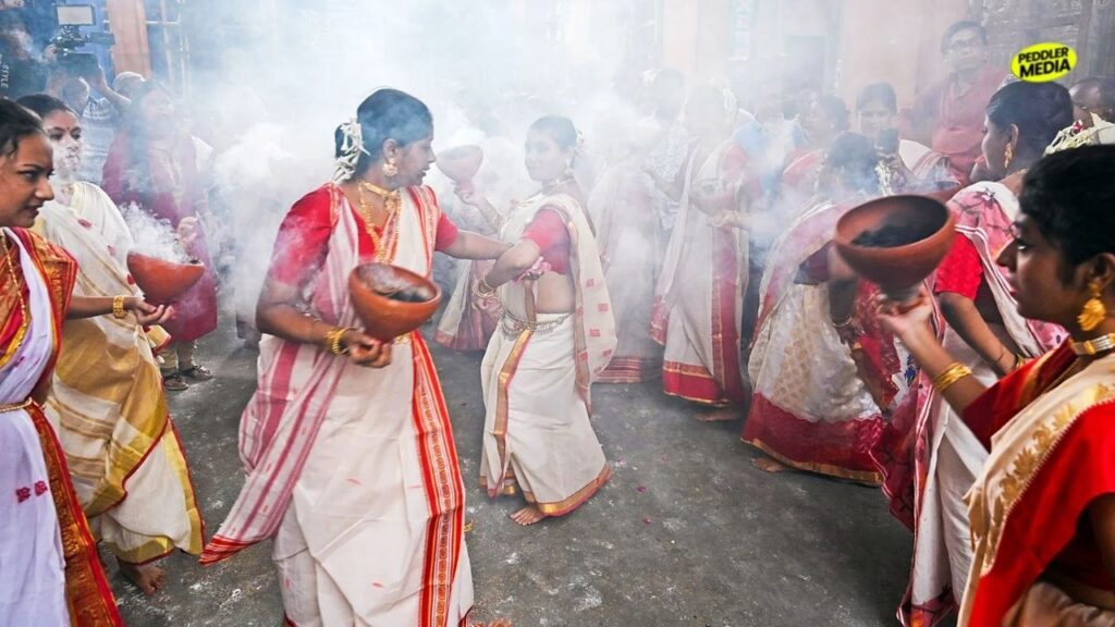 Bengali women wearing traditional white sarees with red borders performing the traditional dhunuchi dance amidst smoke during a festival celebration.