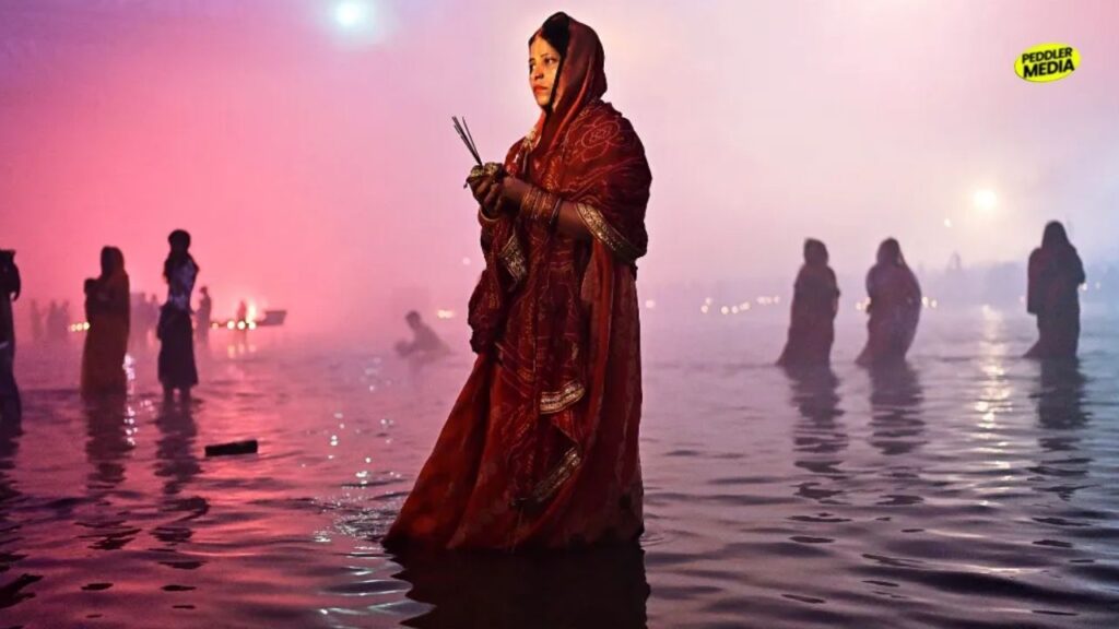 A woman in a traditional red saree standing in a river or pond holding burning incense sticks during evening rituals, representing the vibrant festive traditions and Bengali culture.