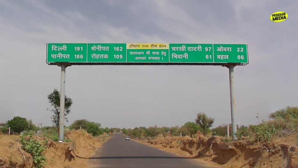 A state highway SH-12 border signboard to explore Haryana showing road distances from Behal and Obra toward Rohtak and Bhiwani.