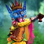 A performer wearing a traditional fierce blue deity mask and vibrant attire holding a smoking dhunuchi burner during a cultural event, representing the Bengali community's traditions.
